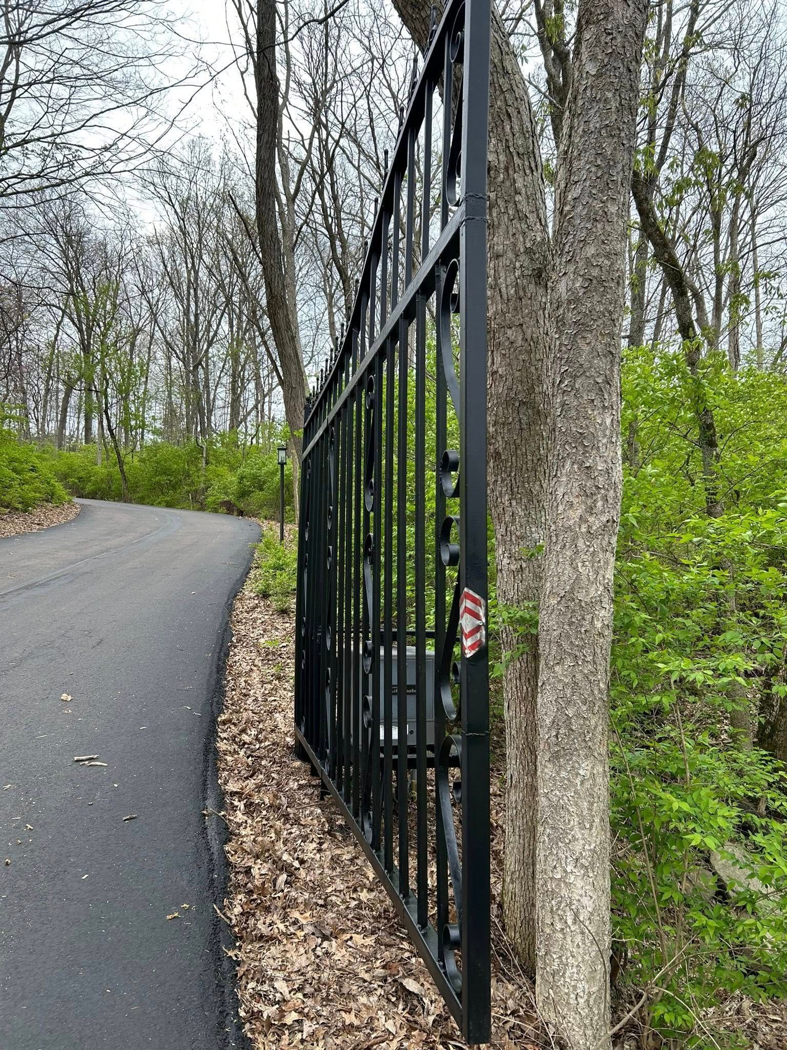 Black metal gate next to a tree and a winding paved road, set in a wooded area.