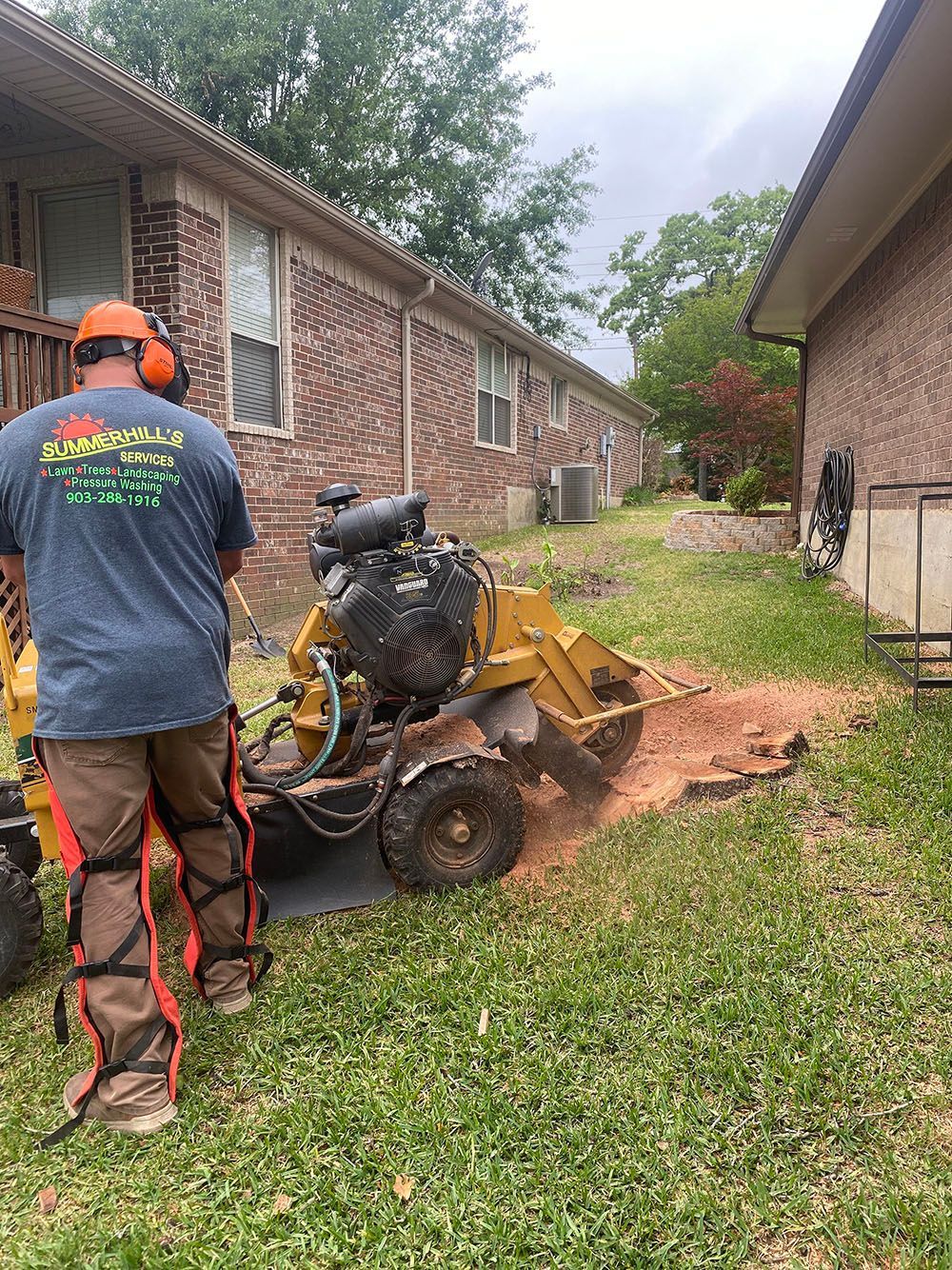 Worker operating a yellow trenching machine beside a brick house, digging a narrow path in the grass