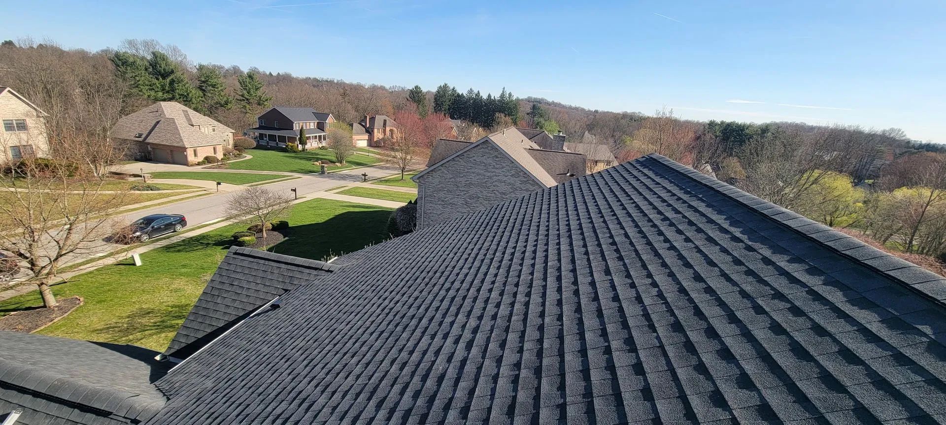 A high-angle view of a dark, textured shingle roof overlooking a suburban street with houses and trees.