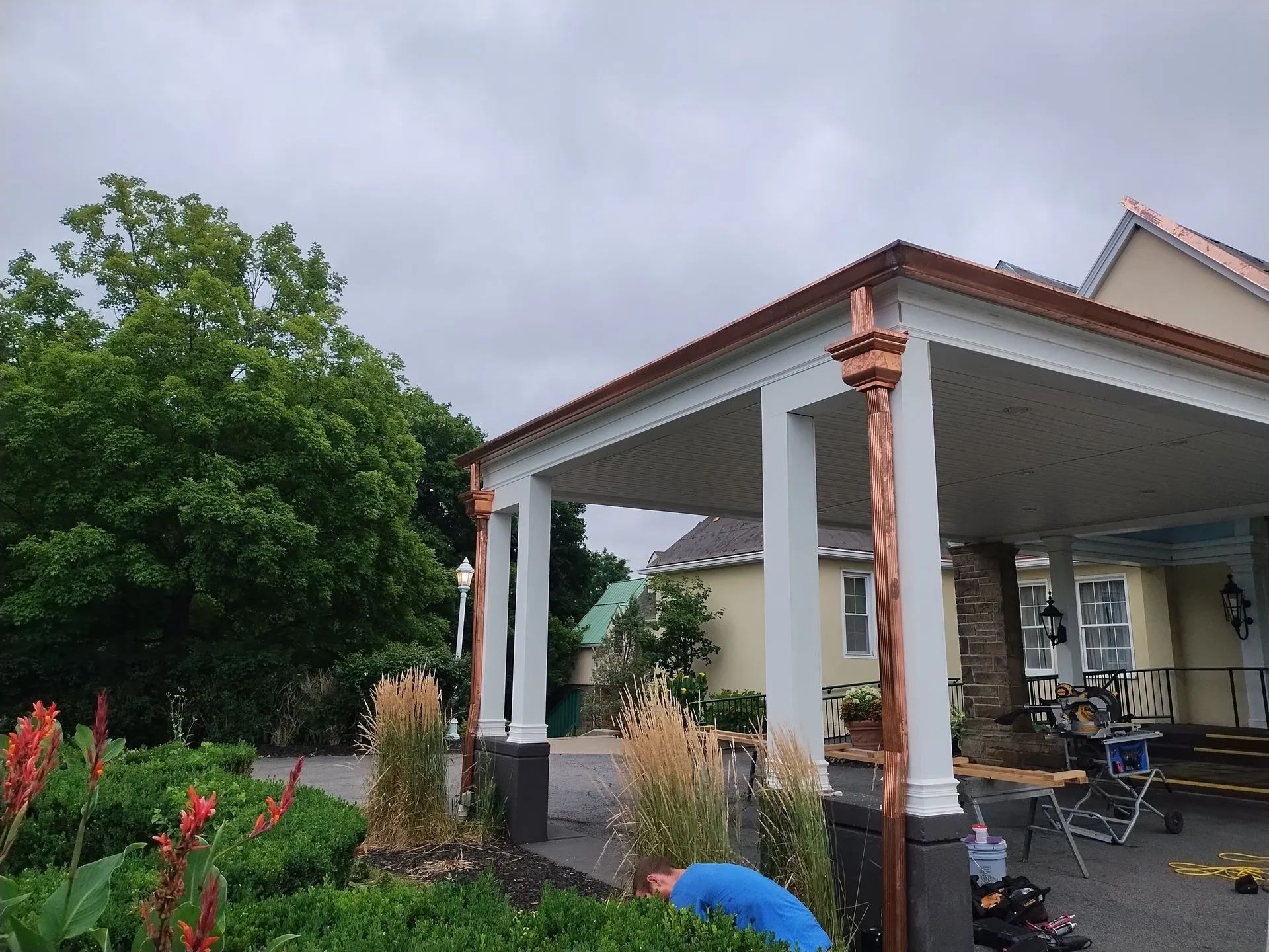 A carport with white pillars and copper accents stands in front of a house, with greenery and a person nearby.