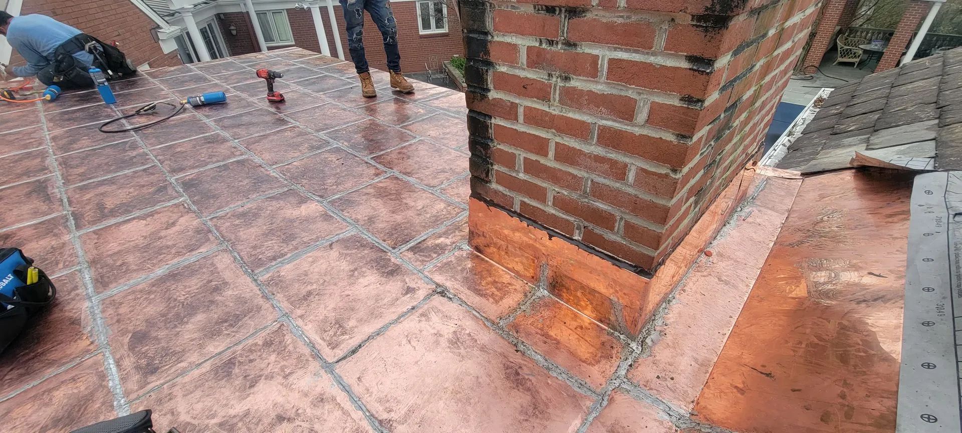 Workers install copper roof flashing and sheets around a brick chimney on a house roof.