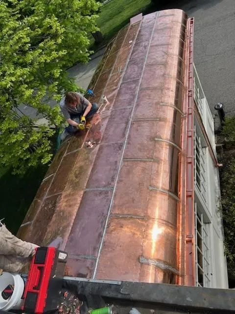 A person working on a copper-paneled roof, soldering metal seams, next to green trees.