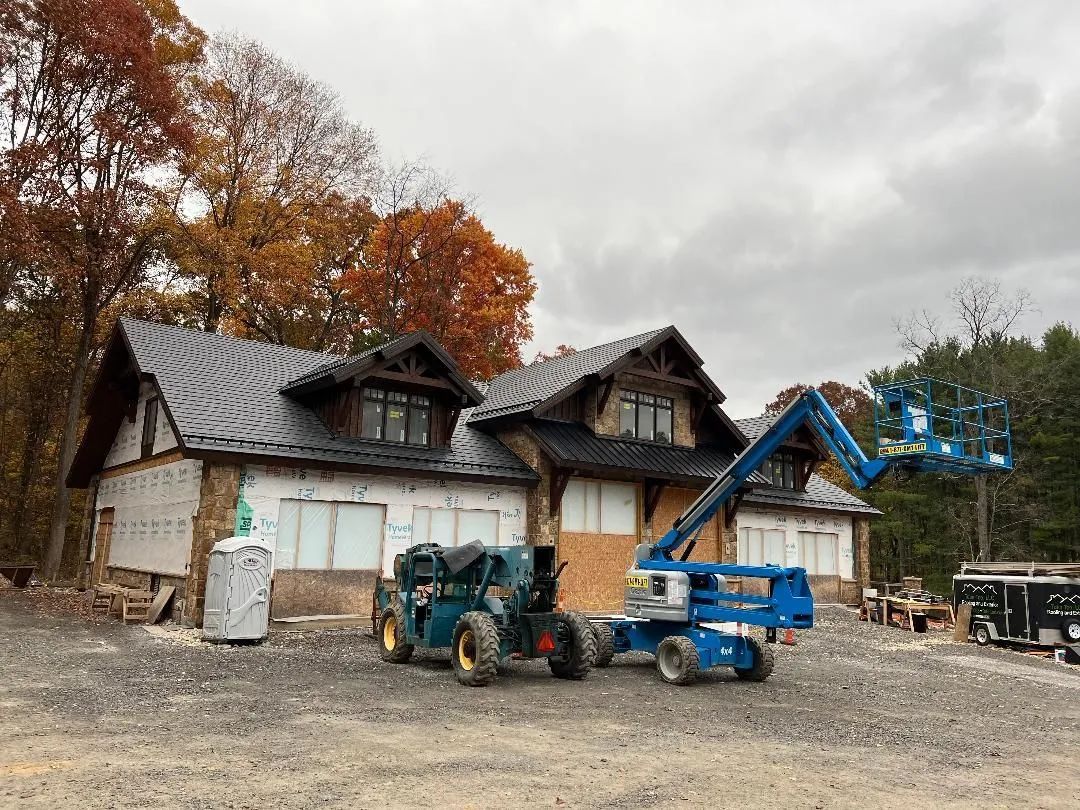 A house under construction with a dark metal roof, two construction vehicles, and a portable toilet on a gravel lot.