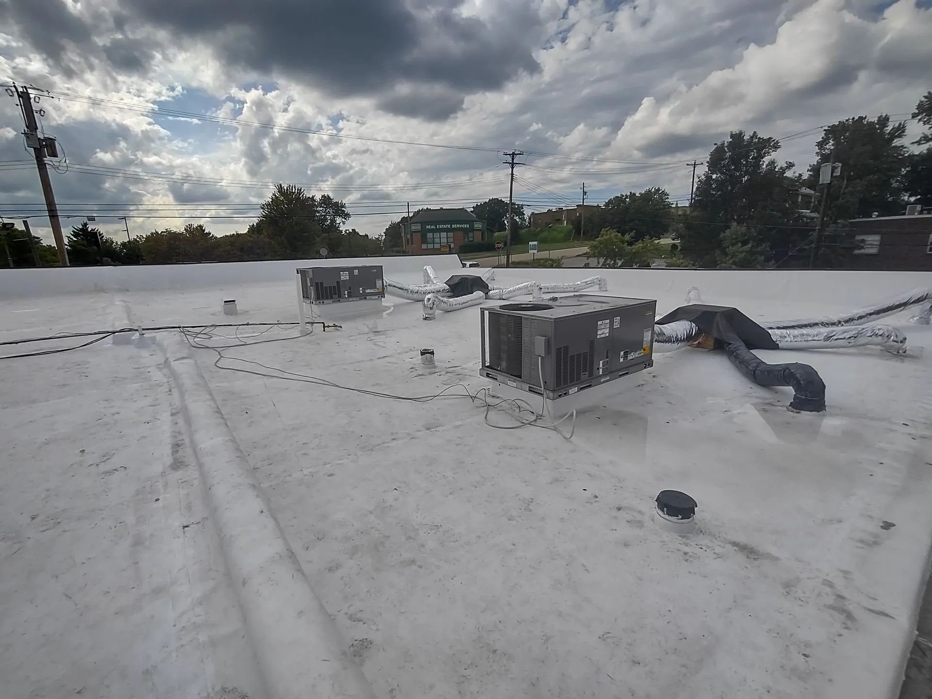 A flat, white commercial roof under a cloudy sky featuring two HVAC units and connecting ventilation ductwork.