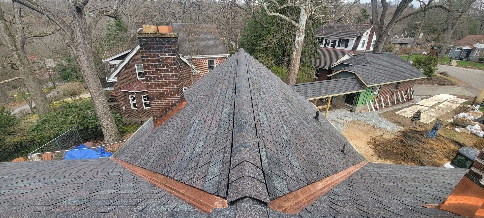A high-angle view of a grey shingled roof under construction, featuring a copper flashing detail and nearby houses.