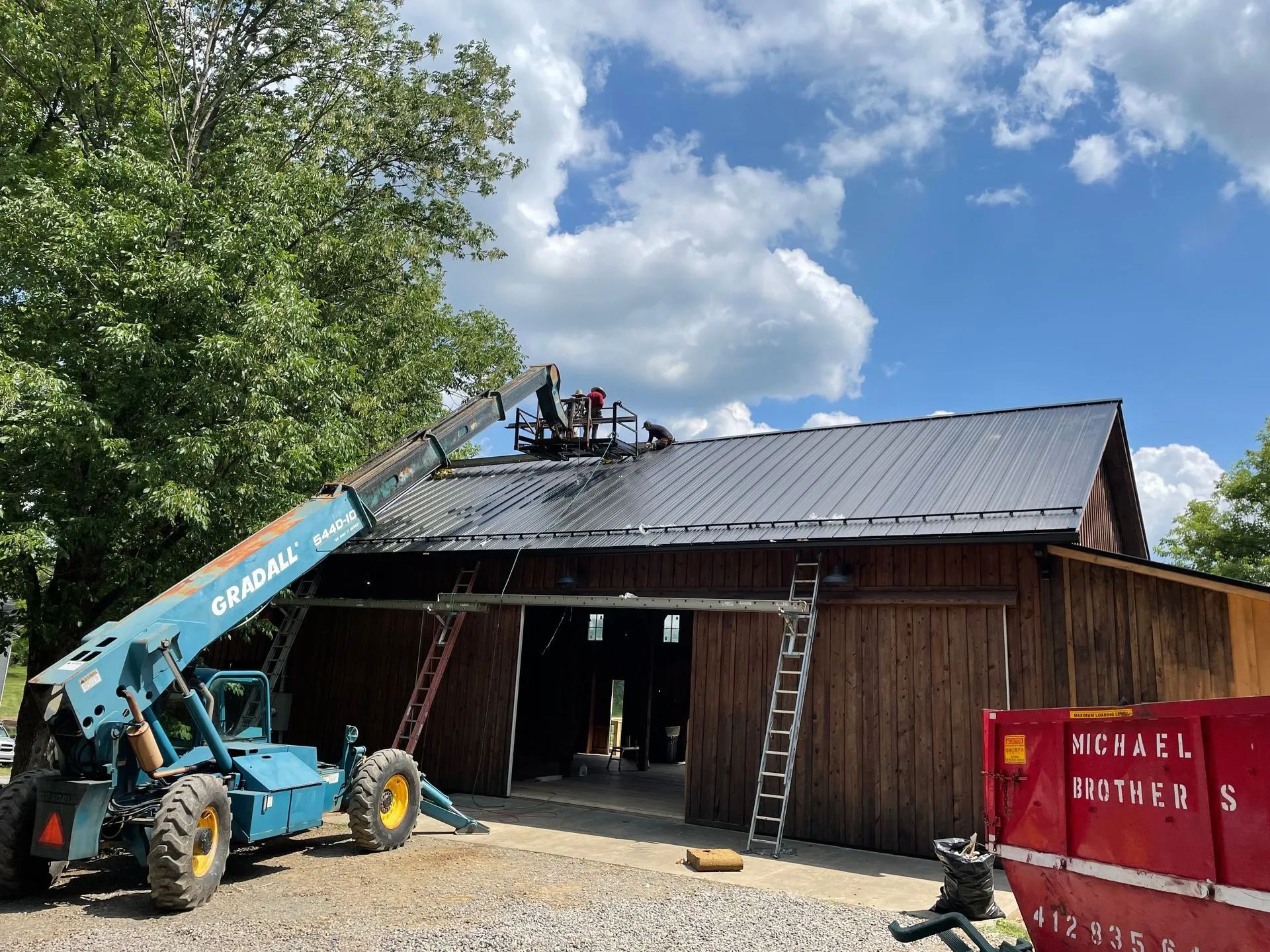 Workers on a lift platform install a black metal roof on a rustic brown barn with a blue telehandler nearby.