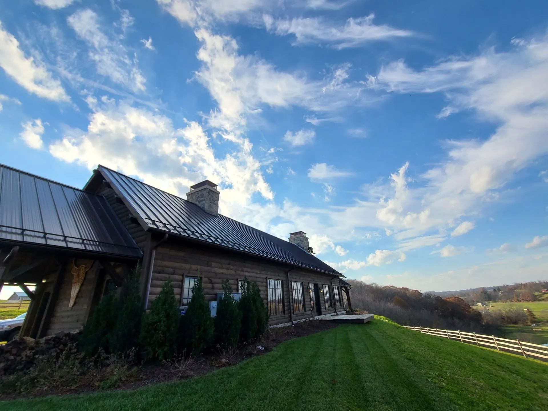A rustic log cabin with a dark metal roof under a bright, cloudy blue sky, situated on a grassy hill overlooking a valley.