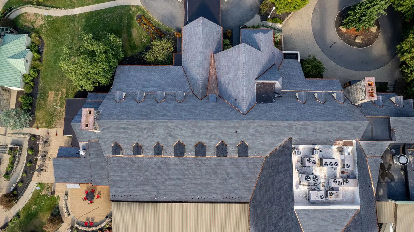 Aerial view of a large building with a dark shingled roof, multiple chimneys, and a flat section with rooftop equipment.