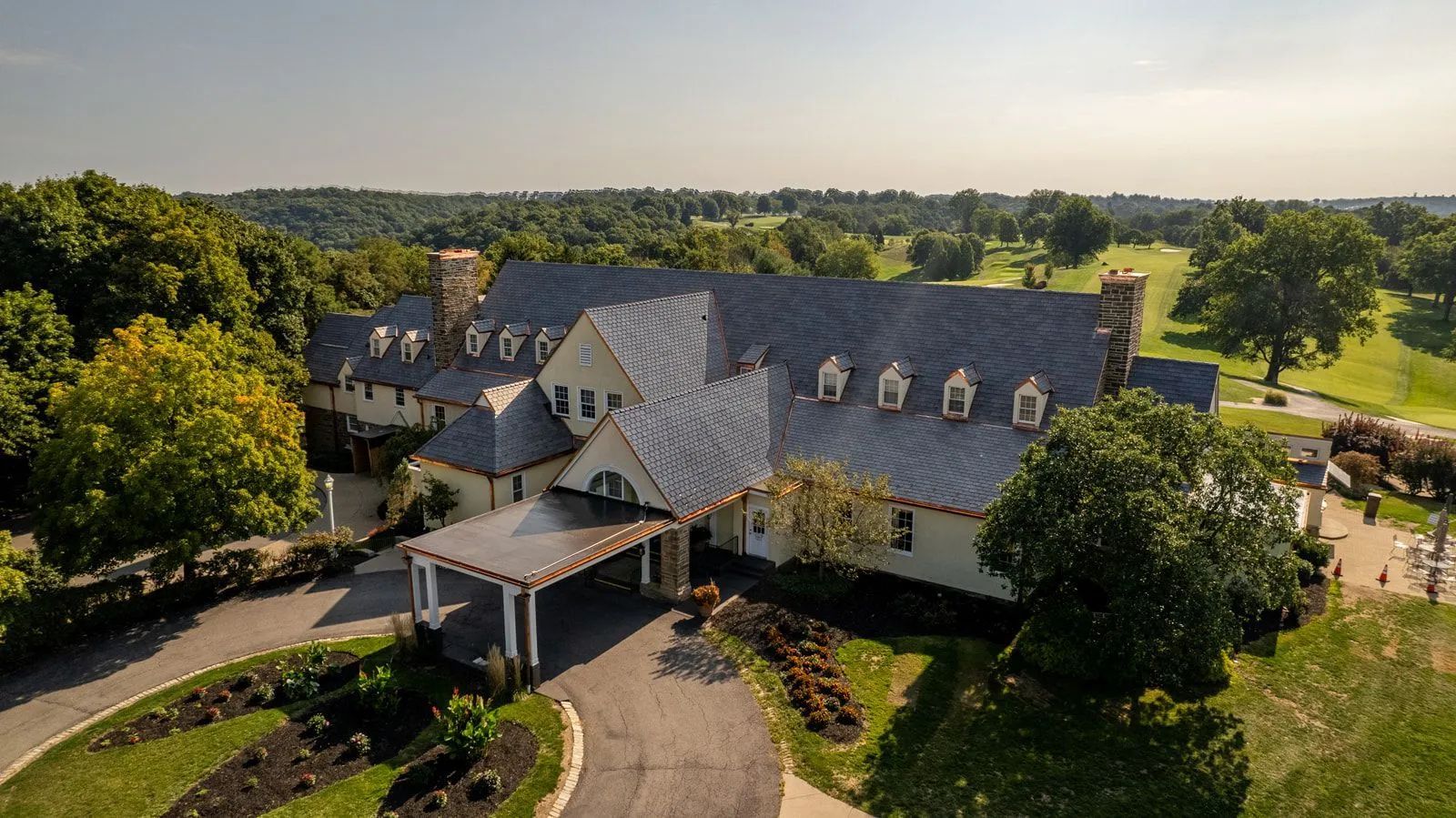 An aerial view of a stone country club building with a slate roof, a portico entrance, and surrounding green golf course.