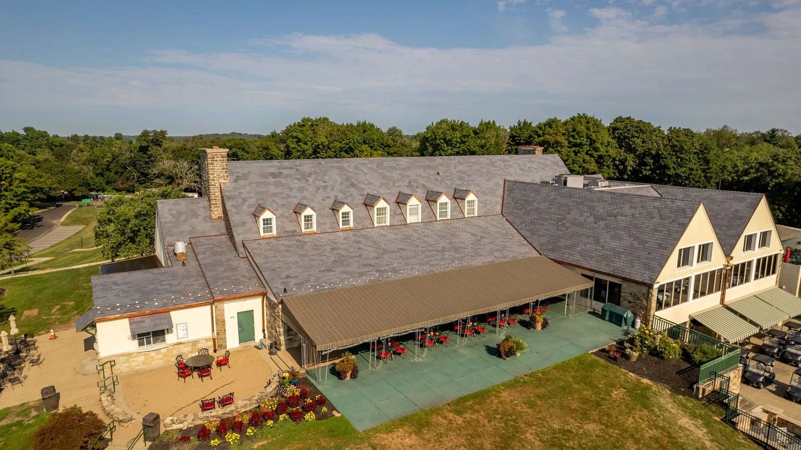 A large stone building with a shingled roof, a green patio, and surrounding trees under a bright blue sky.