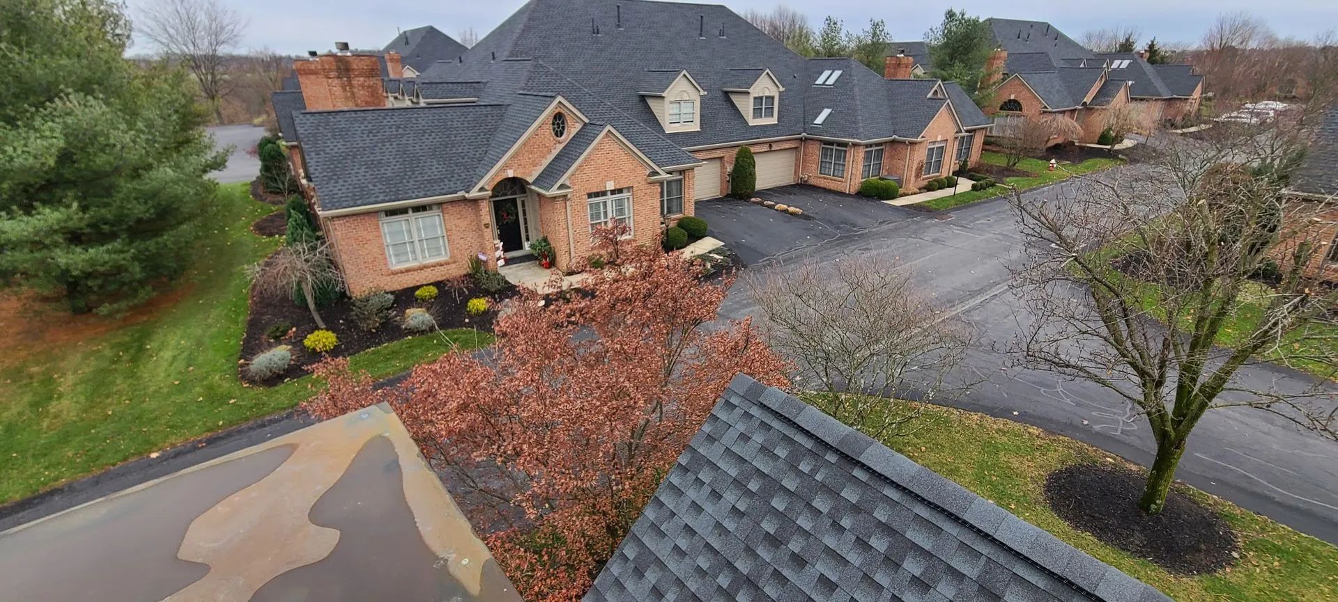An elevated, wide-angle view of a brick house with a dark roof and a paved driveway, surrounded by trees during autumn.