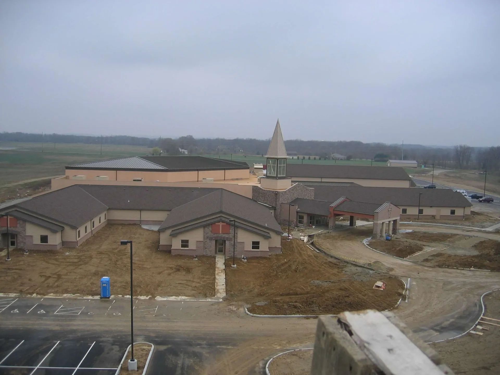 A large, tan building under construction with a steeple, surrounded by dirt lots and a partially paved parking area.