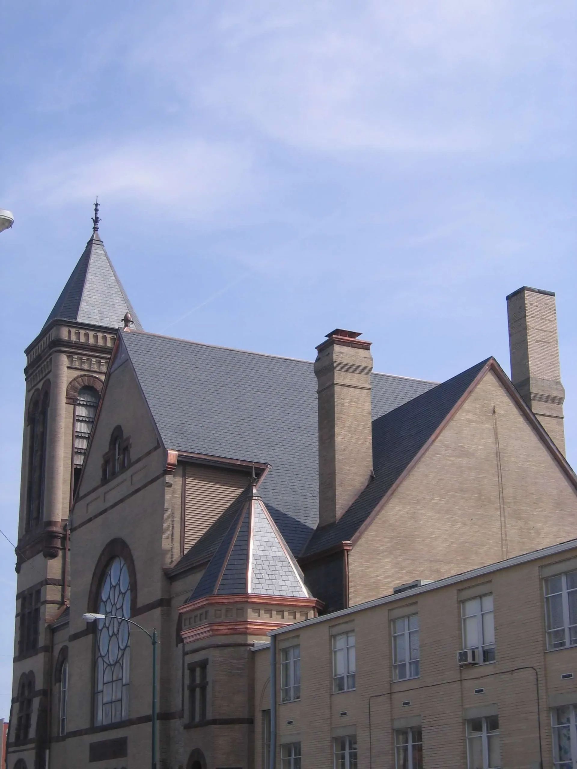 A historic brick church with a tall steeple, a large rose window, and a gray shingled roof under a clear blue sky.