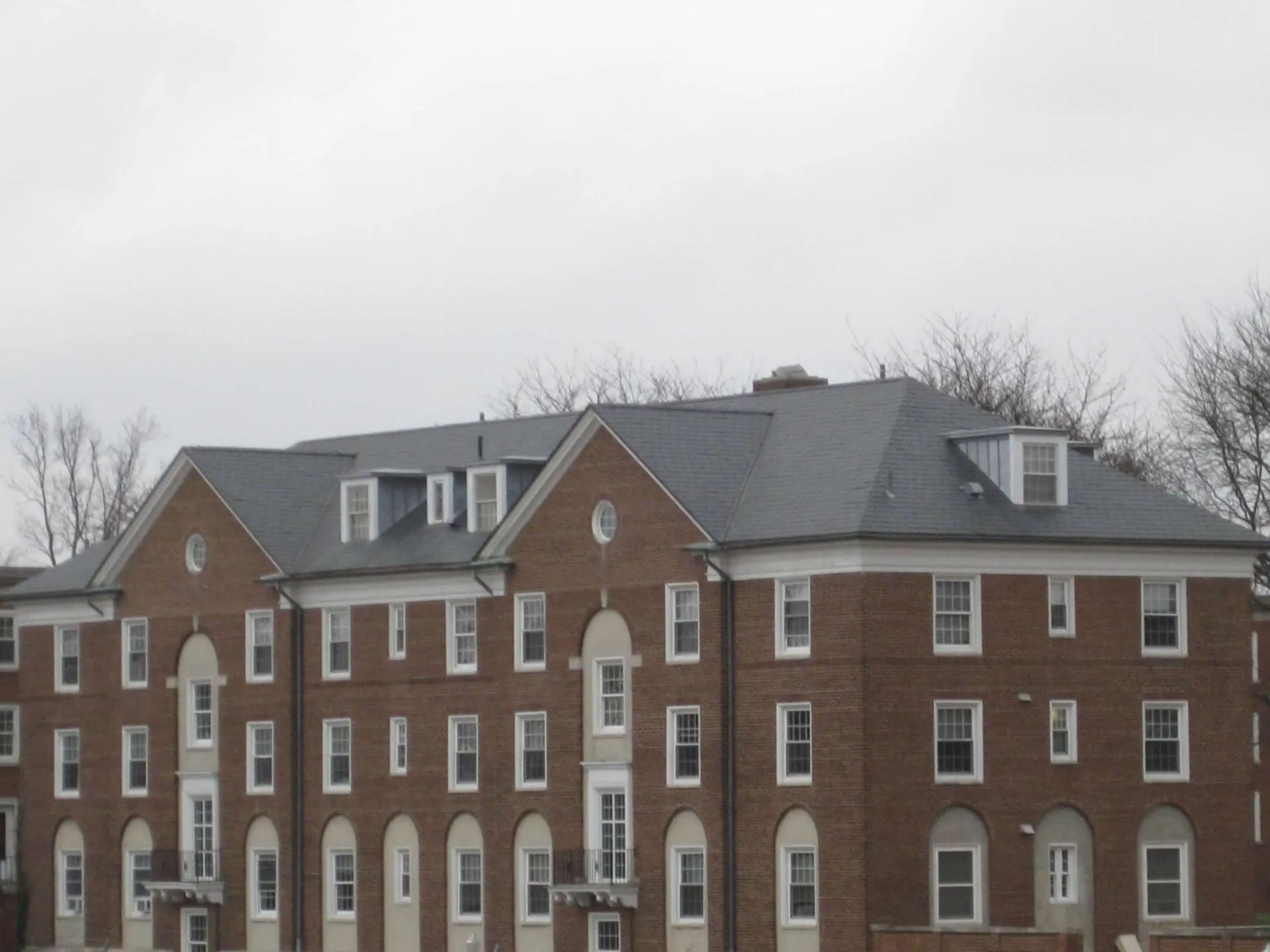 A three-story, red brick residential building with a dark shingled roof, multiple dormer windows, and arched entryways.