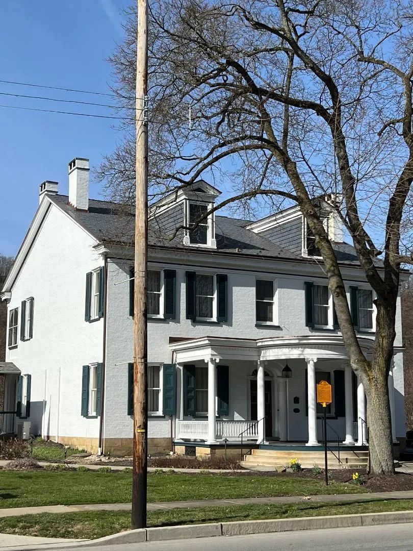 A two-story, white brick house with dark shutters, a porch, and dormer windows under a clear blue sky.