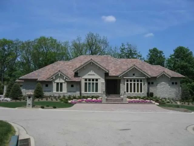 Single-story stone house with a light-colored roof, surrounded by trees and a paved driveway under a blue sky.