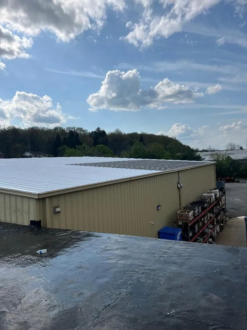 A tan warehouse with a white, partially sunlit metal roof under a blue sky with white clouds, next to a rack of supplies.