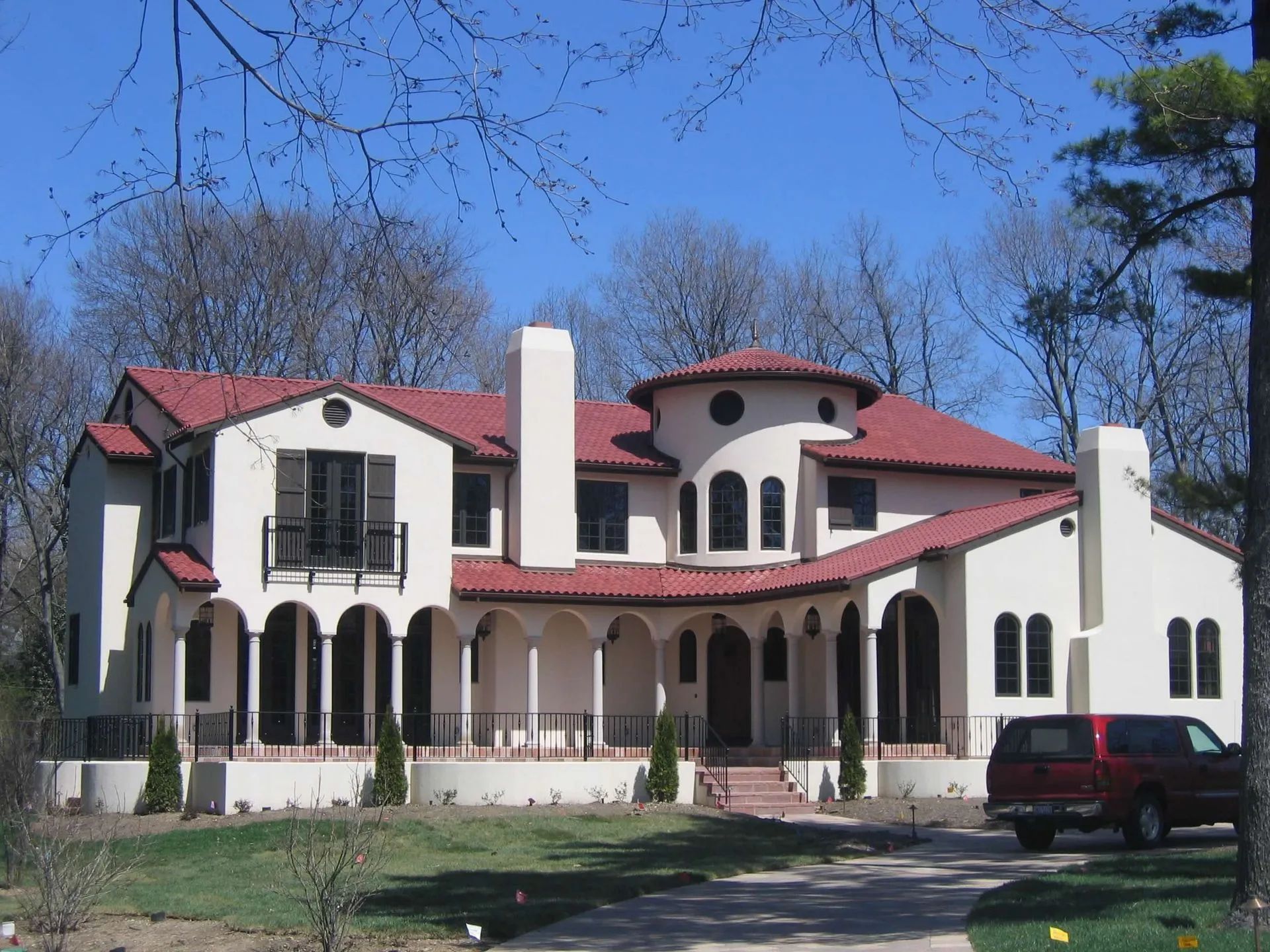 A two-story stucco Mediterranean-style house with a red tile roof, arched porch, and a circular tower, set among trees.