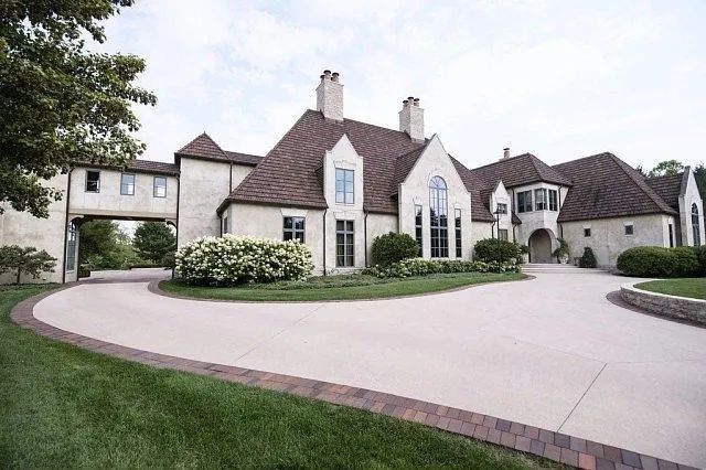A large, two-story stone house with a steep brown roof and a wide, curved concrete driveway.