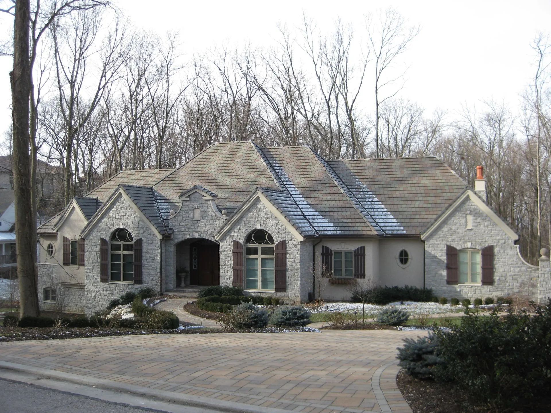 A single-story, stone-faced house with a gray roof and dark brown shutters, nestled in a wooded setting.