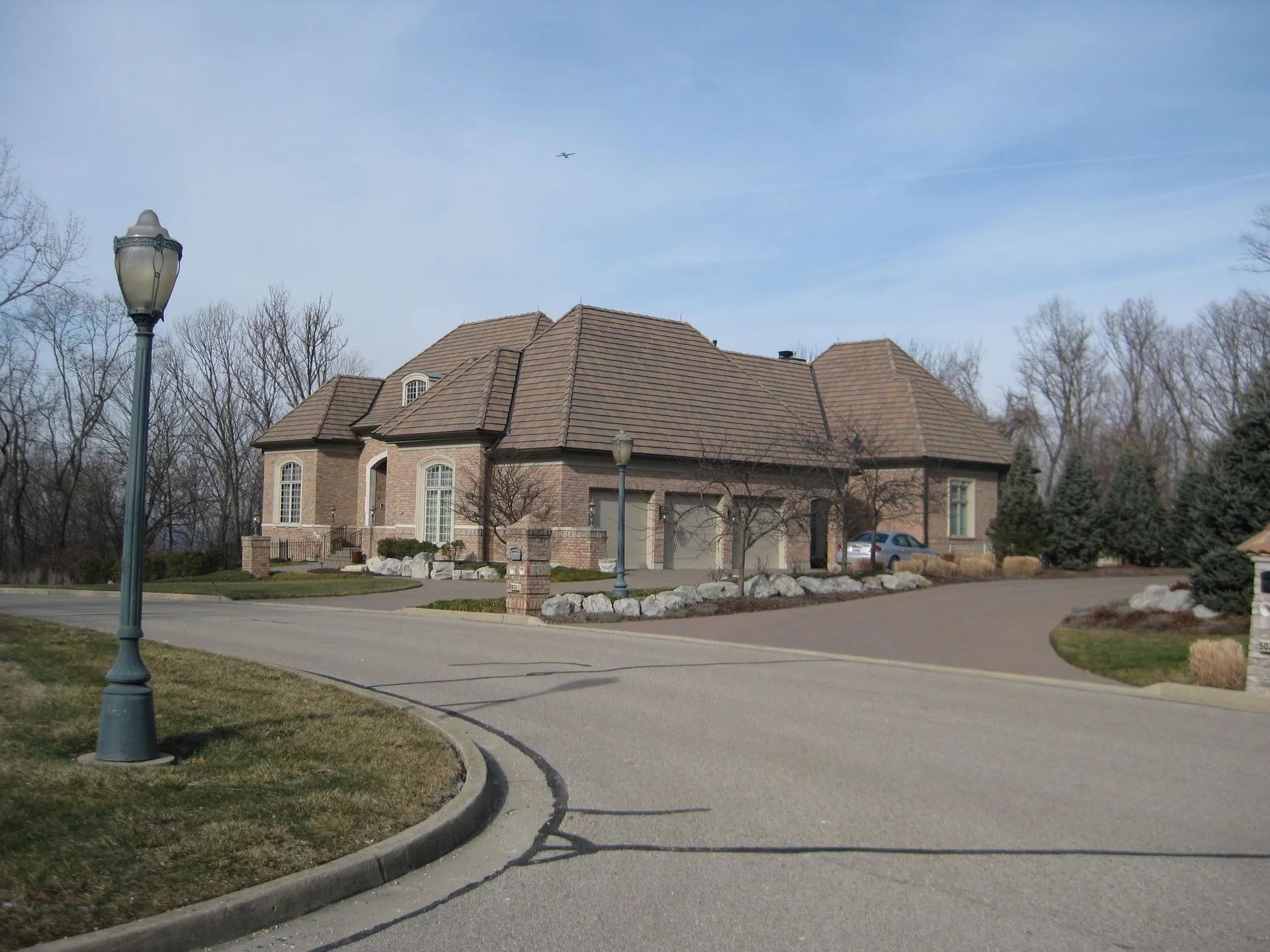 A large, tan brick house with a multi-gabled roof and a three-car garage, viewed from a curved driveway with a lamp post.