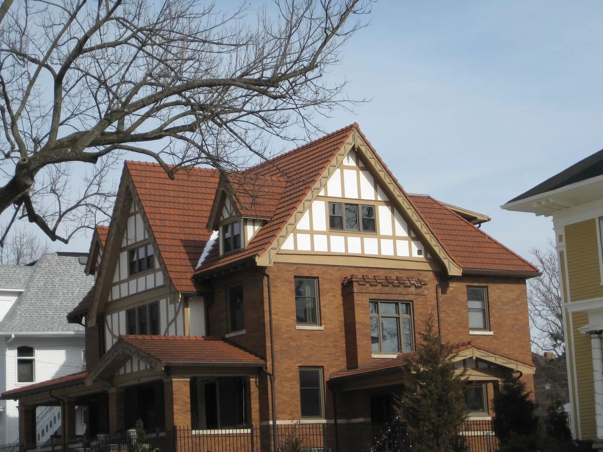 A two-story Tudor Revival home with red-brown brick, timber framing, and a reddish-brown shingled roof under a clear sky.
