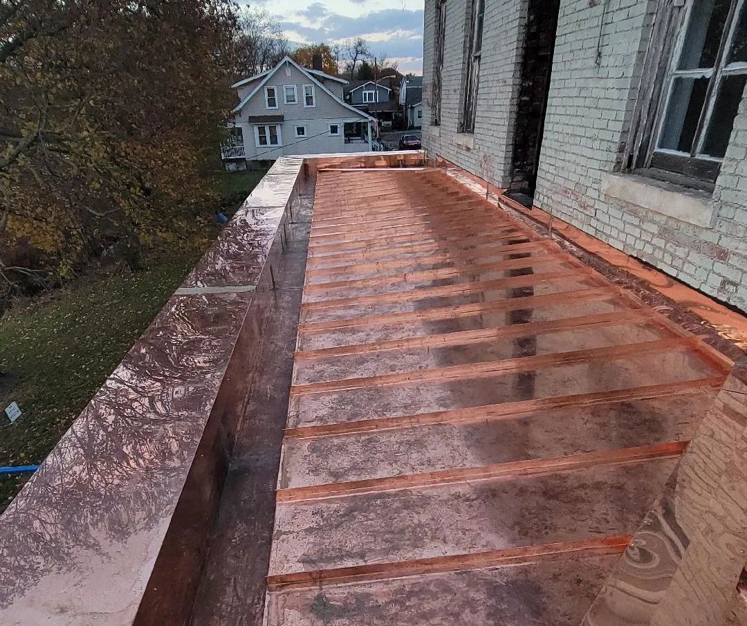 A high-angle view of a newly installed copper roof with prominent standing seams, attached to a light-colored brick wall.