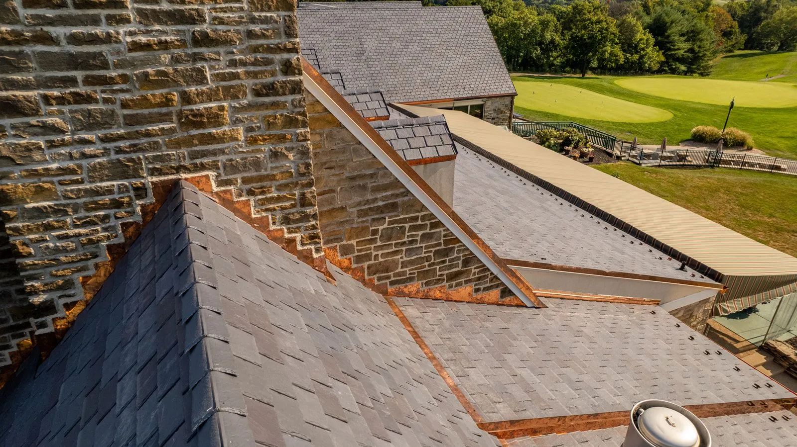A high-angle view of a stone building with a dark shingled roof, copper flashing, and a green landscape in the background.