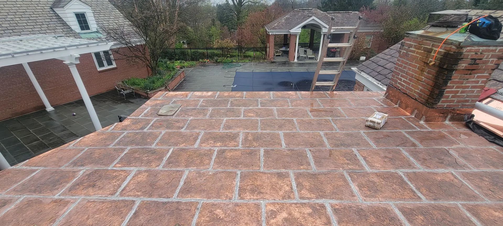 A high-angle view of a brick-tiled flat roof, with a brick chimney and a wooden ladder leaning against a background house.