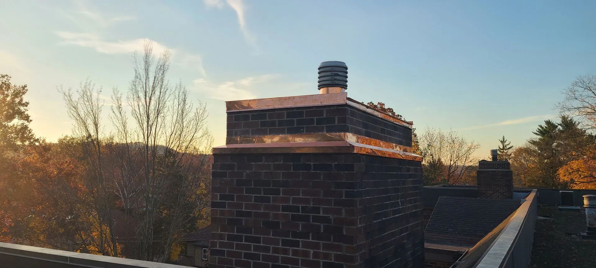 A dark brick chimney with a copper cap and a metal flue on a roof, set against a bright, golden-hour sky.