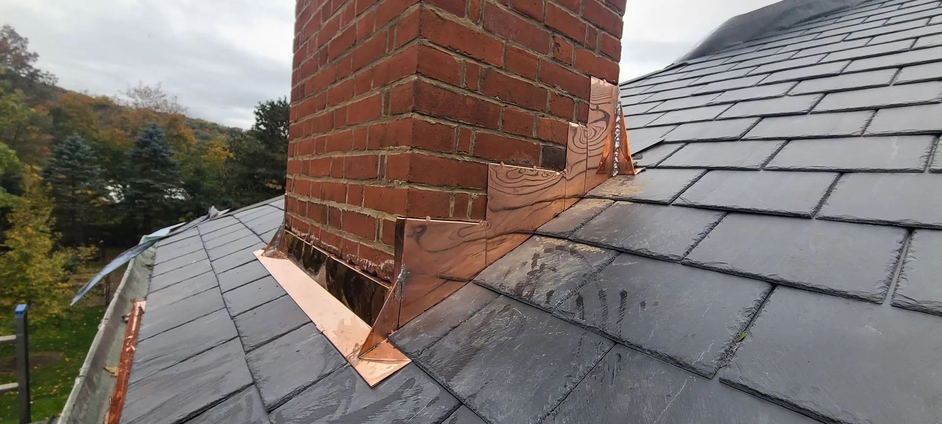 Copper flashing installed around a brick chimney on a dark slate roof, showing a section of exposed roof structure.