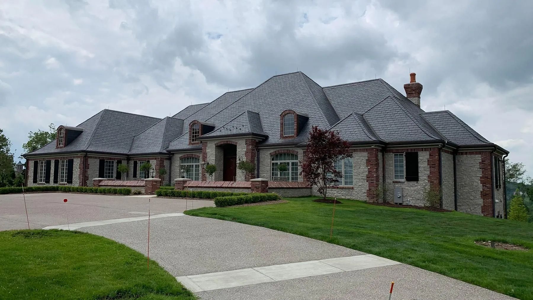 A large, gray stone mansion with a dark shingled roof, set on a green lawn with a gravel driveway under a cloudy sky.