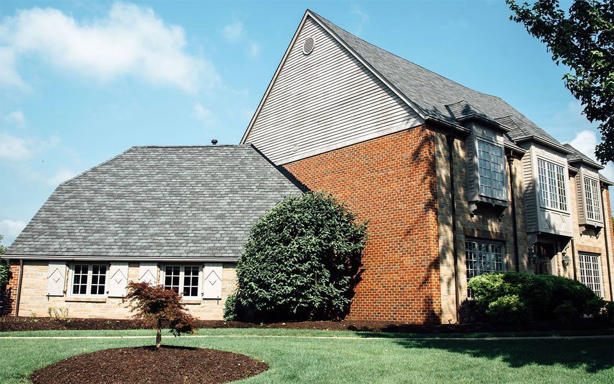 A house with a steep gray shingled roof, red brick siding, and light-colored stone, set against a green lawn and blue sky.