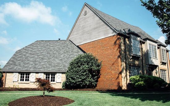 A house with a steep gray shingled roof, red brick siding, and light-colored stone, set against a green lawn and blue sky.