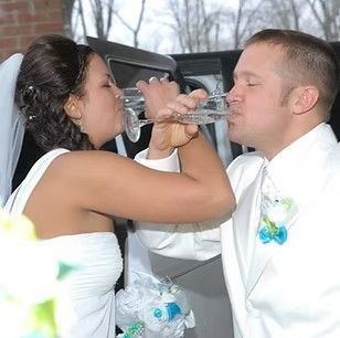 Bride and groom in white wedding attire sharing a drink in a car seat backseat.