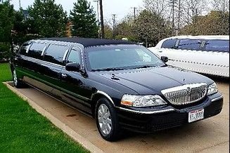 Black stretch limousine parked on a driveway beside a white limousine, with trees in the background
