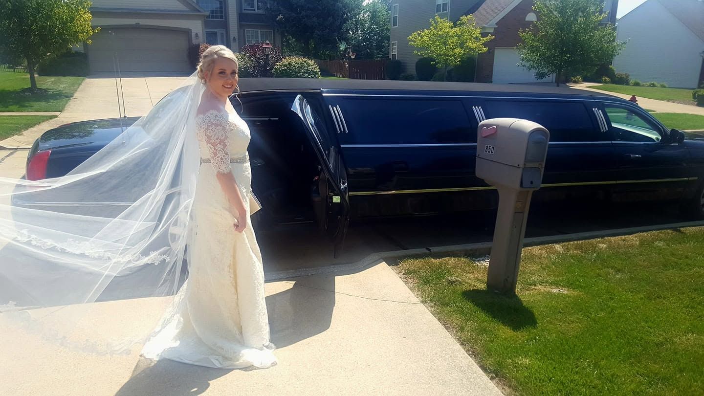 Bride in a white gown and veil walking beside a black limousine on a sunny driveway