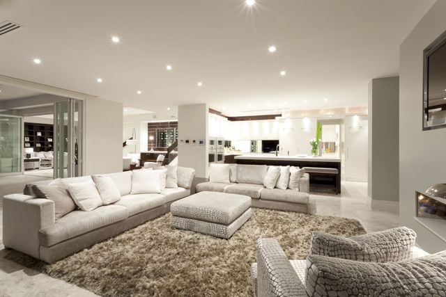 A modern living room with a large rug, couches, and overhead lighting; view into a kitchen.