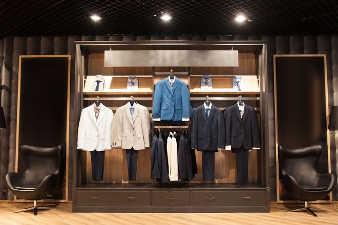 Display of suits in a men's clothing store. Dark wooden shelves with hanging suits, ties, and two black chairs.