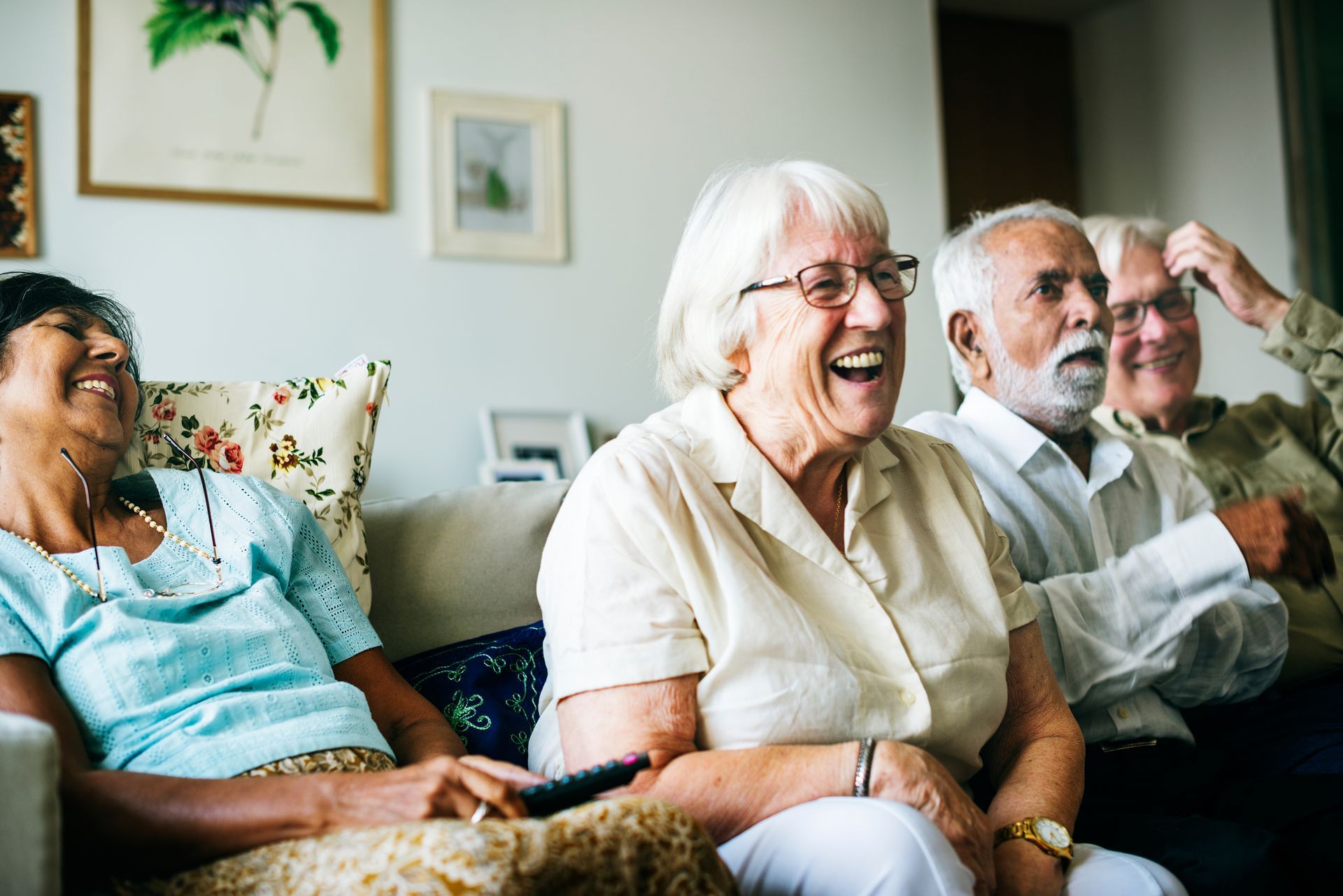 Four older adults laughing on a couch in a living room, some holding a remote.