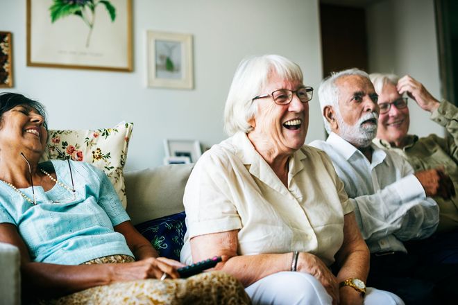 Four older adults laughing on a couch in a living room, some holding a remote.