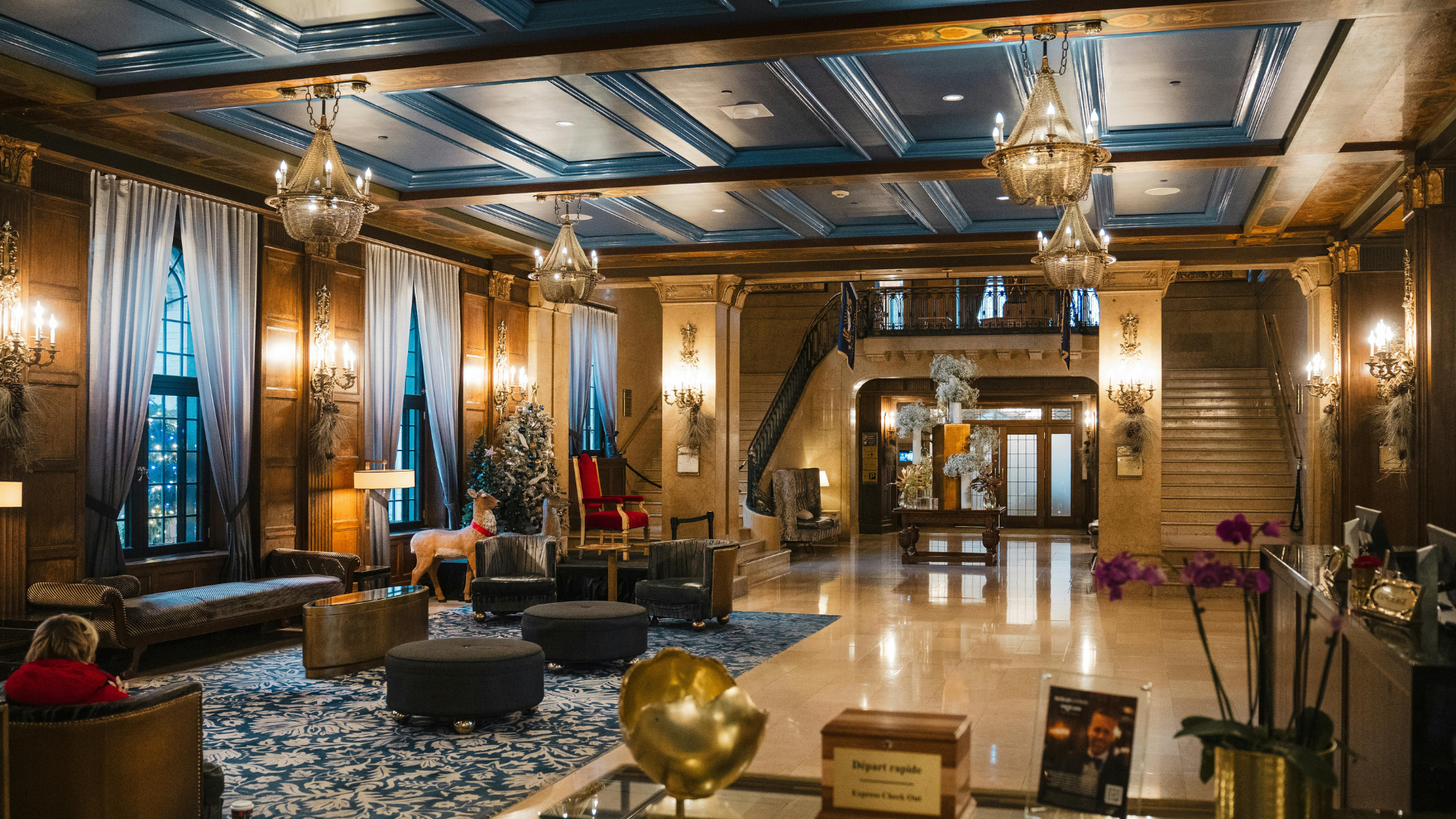 Ornate hotel lobby with chandeliers, marble floors, seating areas, and a grand staircase.
