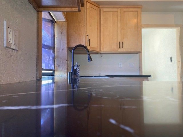 Kitchen with light wood cabinets, dark countertop, and black faucet by a window.