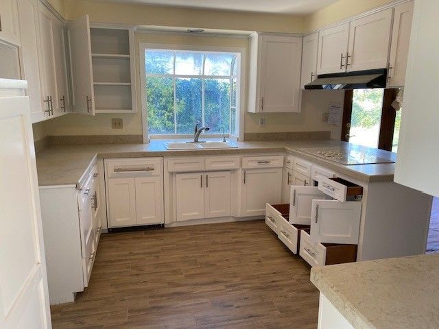 Bright white kitchen with open cabinets, wood floor, and a large window over the sink.