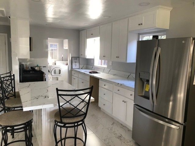 Bright white kitchen with marble countertops, stainless fridge, and black bar stools along the island.