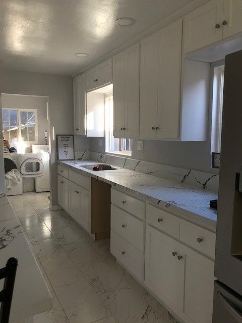Bright white galley kitchen with marble countertops, cabinets, and a window above the sink.