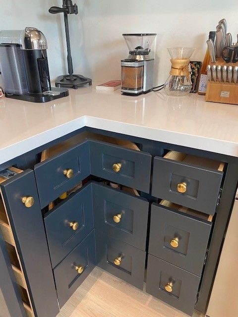 Dark blue kitchen corner cabinet with brass knobs under a white countertop, beside coffee makers and jars