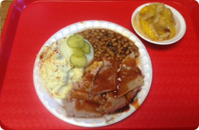 Plate of barbecue: sliced meat, potato salad with pickle, baked beans. Small side of corn. Red tray.