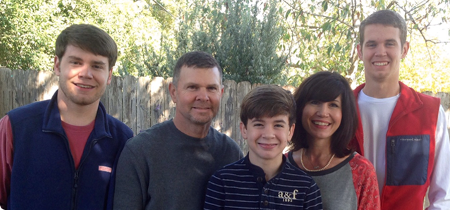 A family of five poses outdoors. Two adult males flank a smiling boy, and a woman and a young man smile nearby.