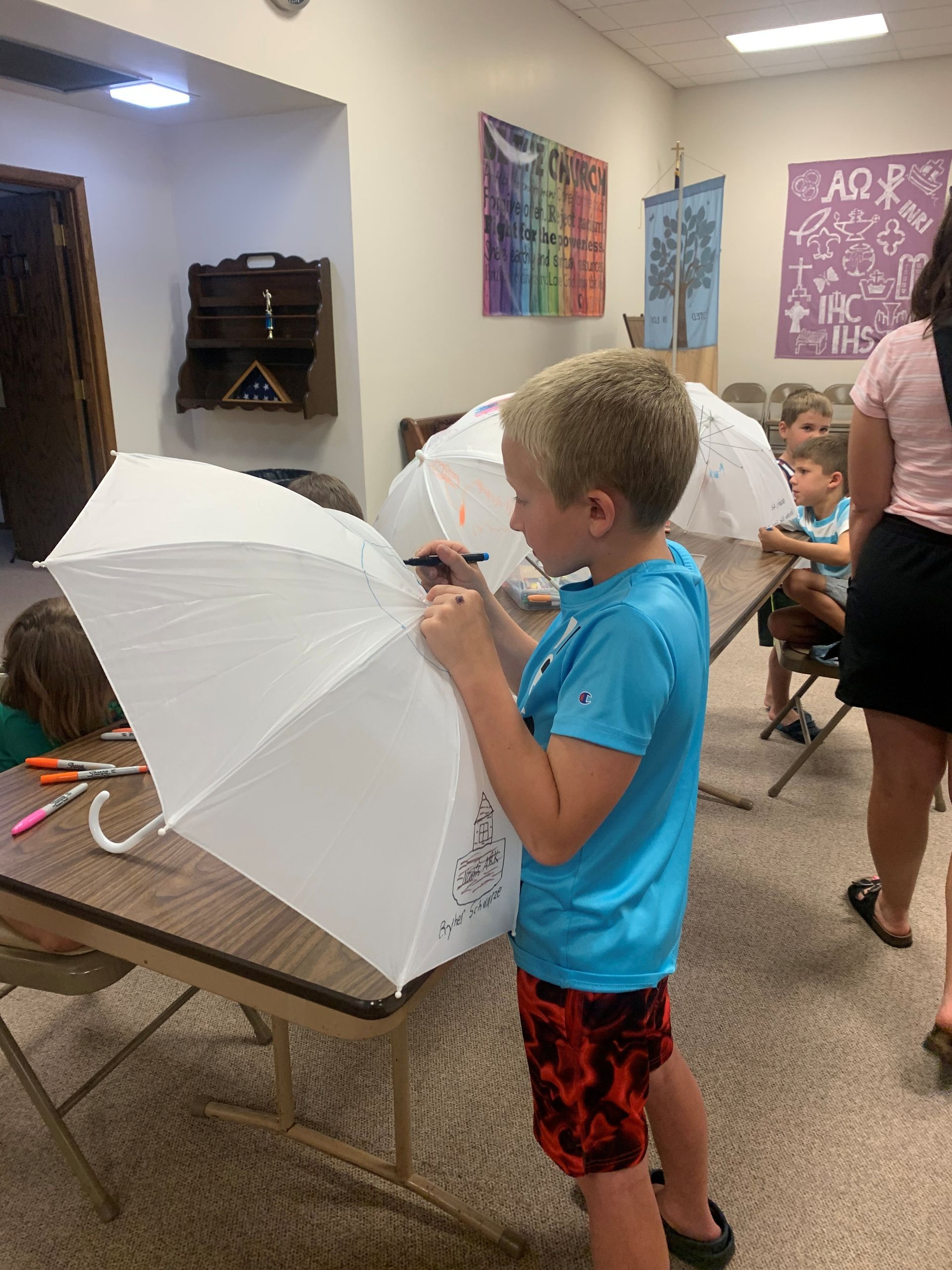 Children making crafts at a table, with one boy holding a white paper umbrella in a classroom.